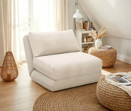 Beiger Schlafsessel mit Kissen auf einem runden Teppich. Auf dem Boden stehen ein geflochtener Hocker und eine Laterne.