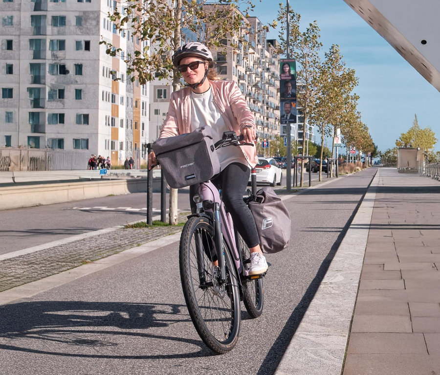 Frau fährt mit einem lila Fahrrad mit einer Fischer Gepäckträgertasche PLUS Cita auf der Vorderseite.