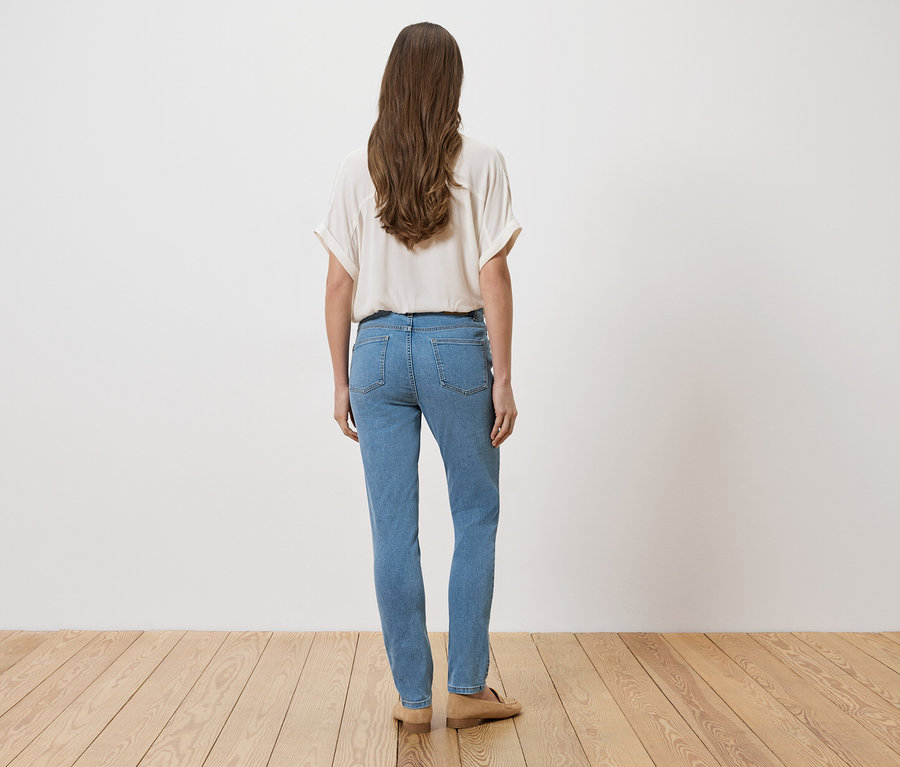 Frau mit langem braunem Haar, von hinten gesehen, trägt eine helle Bluse und blaue Jeans, stehend auf einem Holzboden vor einer weißen Wand.