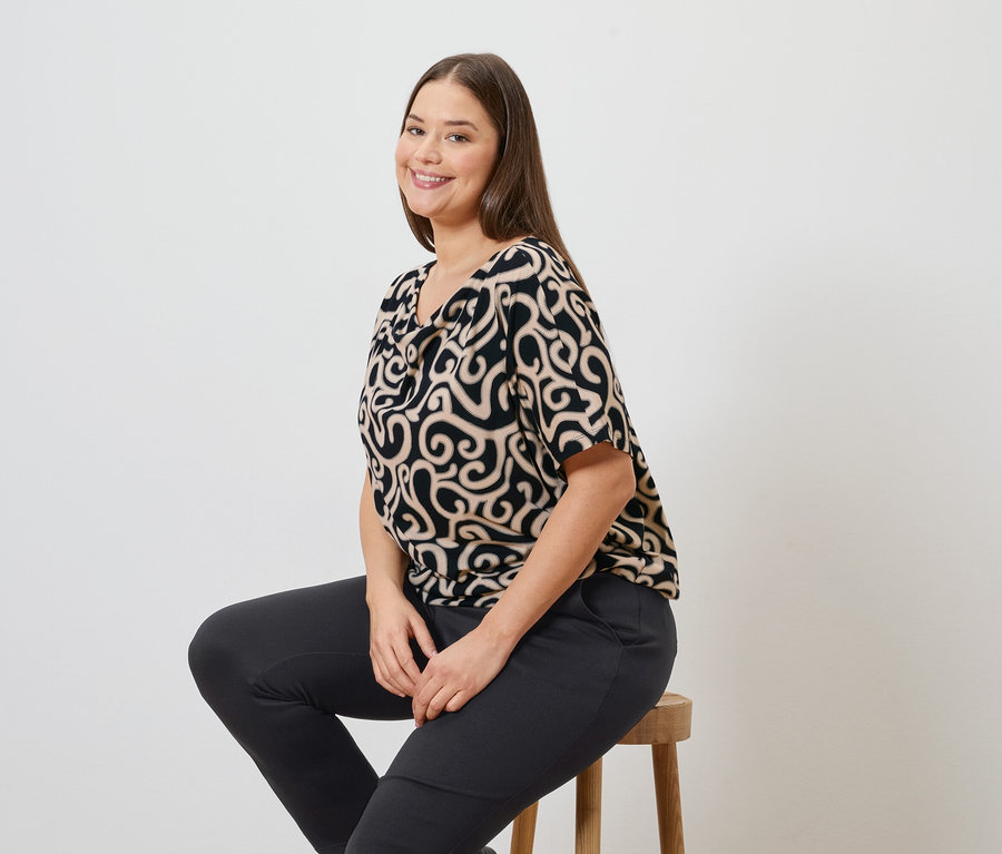 Frau sitzt auf einem Hocker und trägt eine gemusterte Bluse und eine Joggpants.