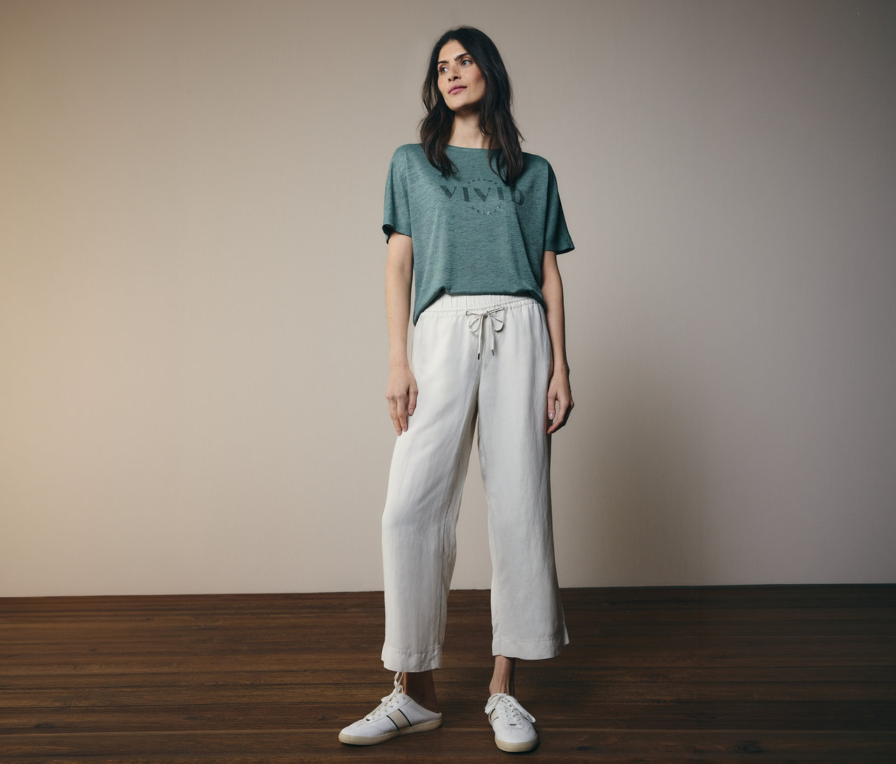 Frau mit dunkelbraunen Haaren, die ein grünes T-Shirt mit der Aufschrift "VIVID" und eine weiße Hose mit Kordelzug und weiße Schuhe trägt.
