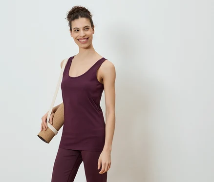 Frau im auberginefarbenen Top und Hose, Yogamatte über der Schulter, lächelt vor einem weißen Hintergrund.