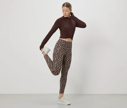 Frau dehnt Bein in braunem Oberteil, Leggings mit Leopardenmuster und weißen Turnschuhen vor einem weißen Hintergrund.