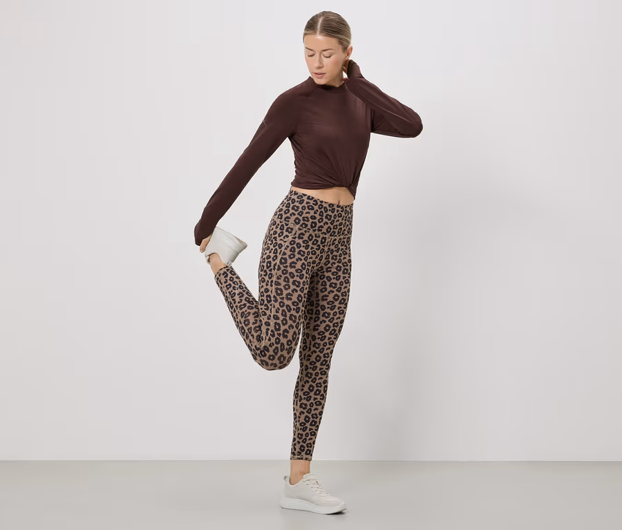 Frau dehnt Bein in braunem Oberteil, Leggings mit Leopardenmuster und weißen Turnschuhen vor einem weißen Hintergrund.