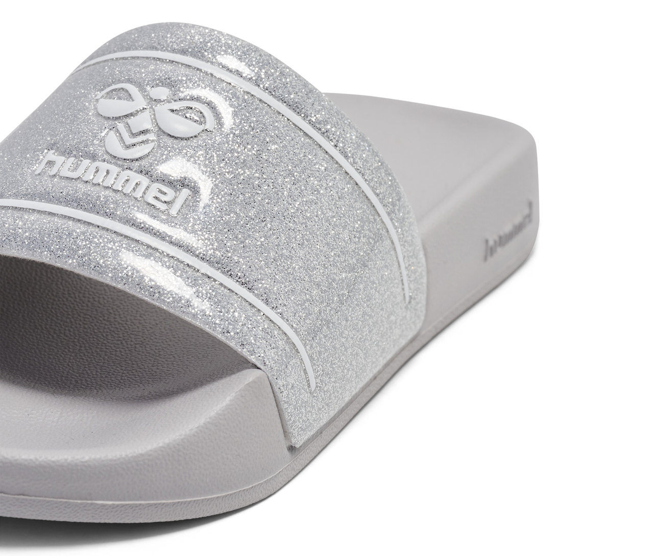 Nahaufnahme von einem hummel® Pool Slide GlitterJR Badelatschen mit silbernem Glitzerriemen und Logo auf grauem Fußbett.