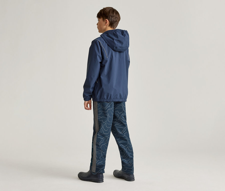 Ein Junge steht mit dem Rücken zur Kamera und trägt eine dunkelblaue Kapuzenjacke mit weißen Streifen, eine dunkelblaue Hose mit hellem Muster und dunkle Stiefel.