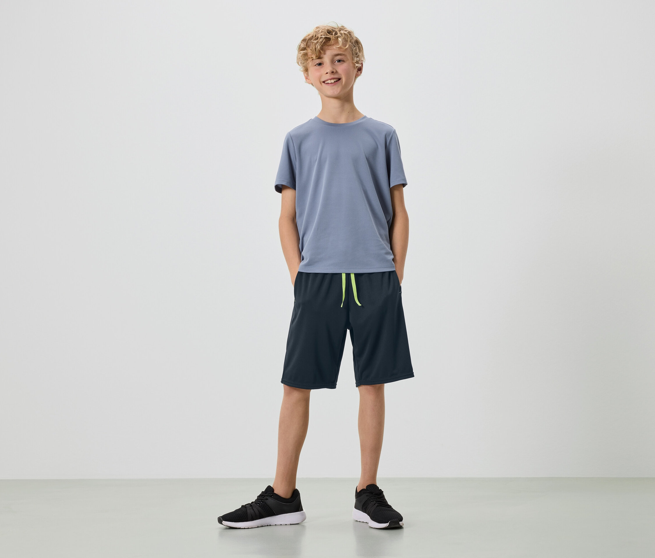 Ein junger Mann mit blonden Locken steht in einem grauen T-Shirt, dunkelblauen Shorts und schwarzen Turnschuhen vor einem weißen Hintergrund.