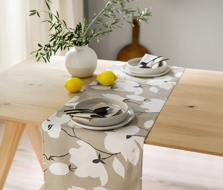 Ein heller Holztisch ist mit einem sandfarbenen SCHÖNER WOHNEN-Kollektion Tischläufer mit weißen Blumenmustern gedeckt. Zwei Gedecke mit je einem Teller, einer Schale, schwarzem Besteck und einer Serviette liegen auf dem Läufer. Eine weiß-beige Vase mit grünen Zweigen und zwei Zitronen stehen ebenfalls auf dem Tisch.