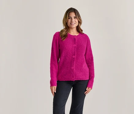 Frau mit braunen Haaren lächelt, trägt eine pinke strukturierte Strickjacke mit Knöpfen und dunkle Jeans vor hellem Hintergrund.