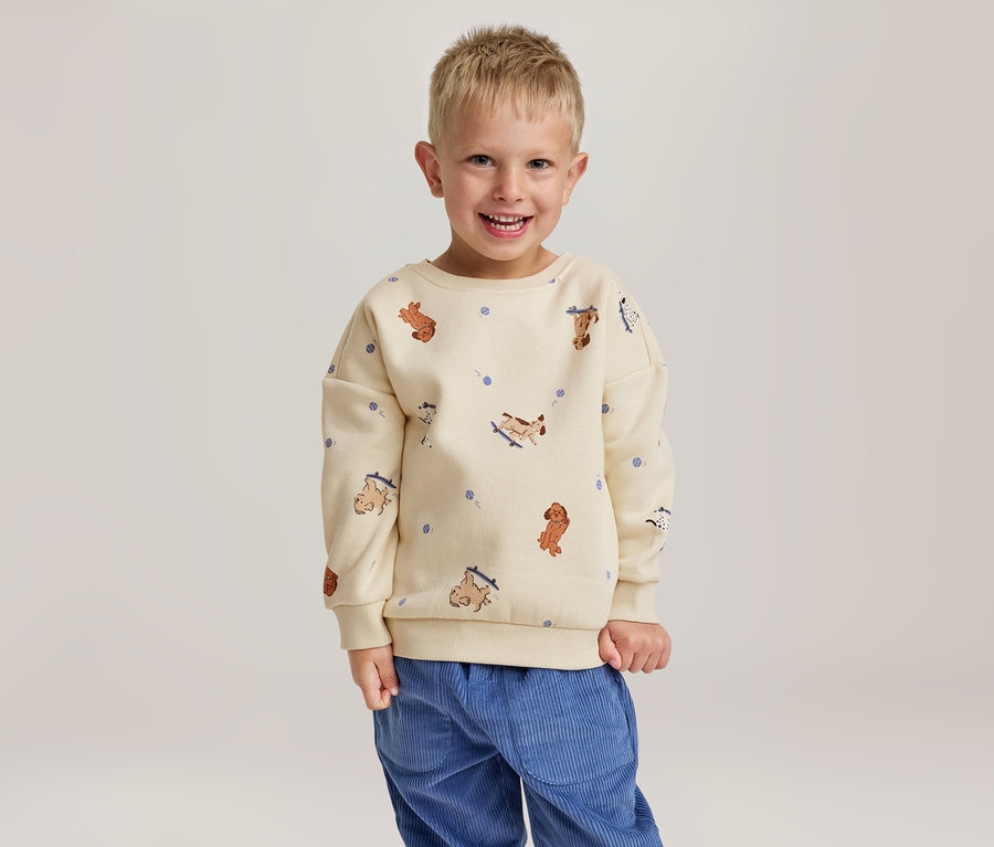 Ein lächelnder kleiner Junge trägt ein cremefarbenes Sweatshirt mit Hundemustern und eine blaue Cordhose.