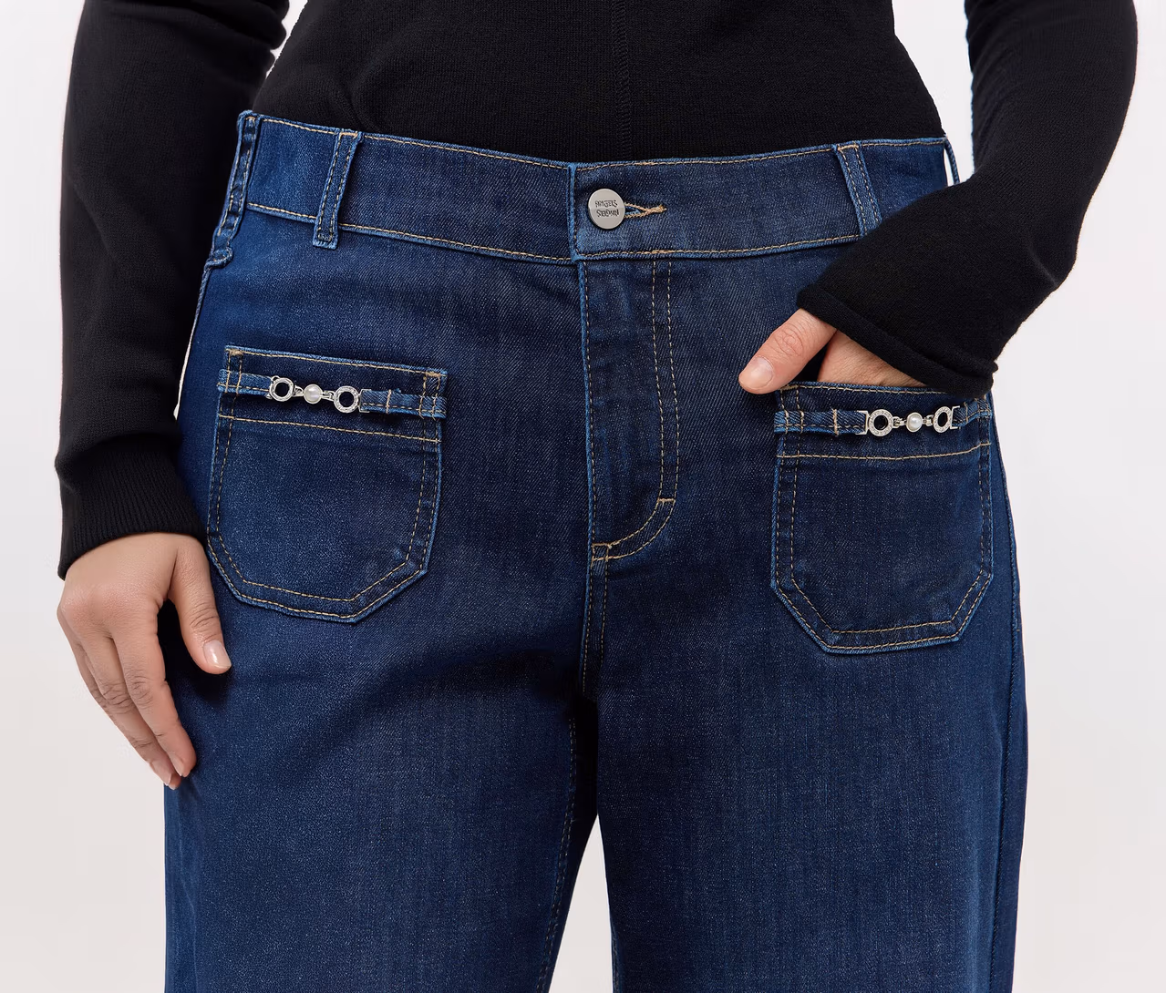 Nahaufnahme einer Person in dunkelblauen Jeans und einem schwarzen Langarmshirt. Eine Hand steckt in der Vordertasche der Jeans, die andere ruht an der Seite. Die Jeans hat dekorative silberfarbene Kettenverzierungen mit Perldetails an den aufgesetzten Taschen.