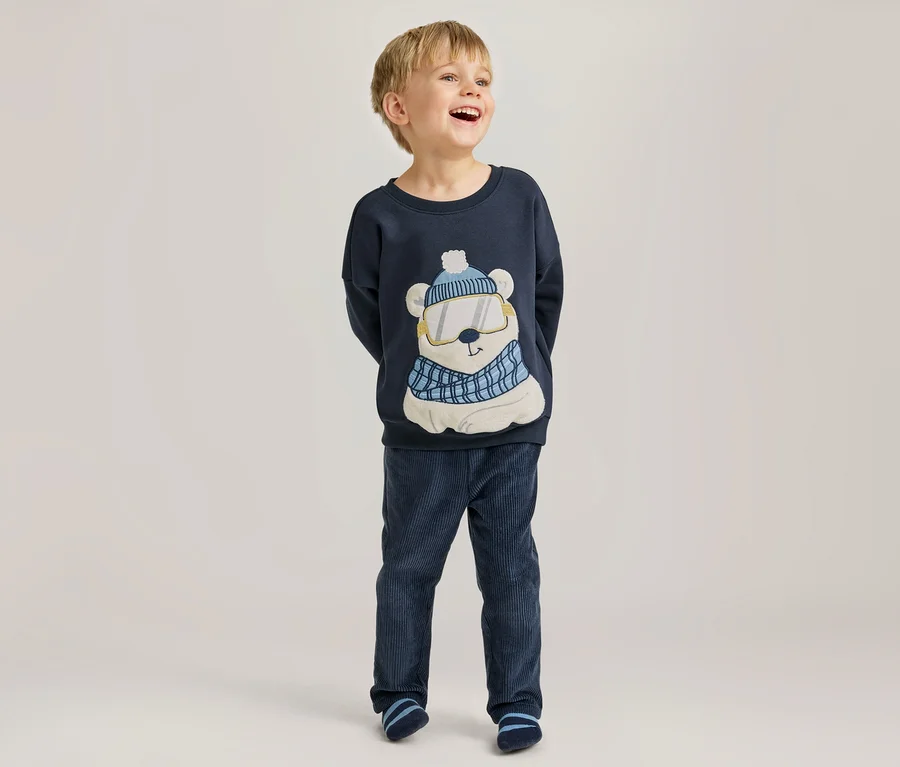 Lachender Junge in einem dunkelblauen Langarmshirt mit Eisbärmotiv, das eine Mütze, Skibrille und einen Schal trägt, dazu dunkelblaue Cordhose und gestreifte Socken.