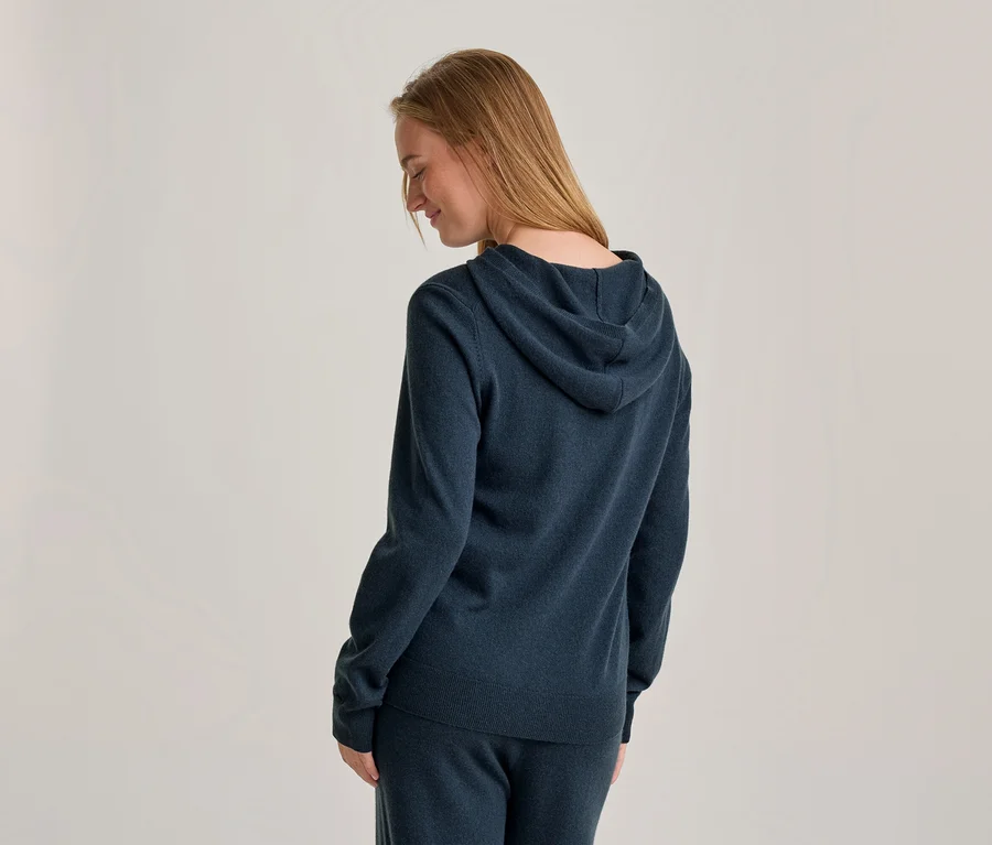 Frau mit langen blonden Haaren, von hinten gesehen, trägt einen dunkelblauen Hoodie und passende Jogginghose, blickt lächelnd nach unten.