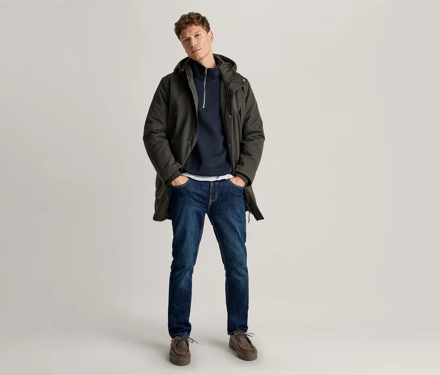 Ein Mann steht mit den Händen in den Taschen, trägt einen dunkelgrünen Kapuzenparka über einem marineblauen Half-Zip-Hoodie, blaue Jeans und braune Schuhe.