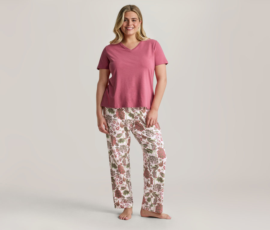 Eine lächelnde Frau steht da und trägt ein pinkes V-Ausschnitt-T-Shirt und weiße Hose mit braunem und pinkem Blumenmuster.