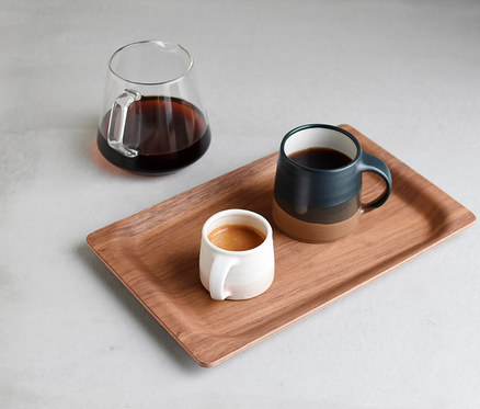 Ein Holztablett mit einer weißen Espressotasse und einem zweifarbigen blau-braunen Keramikbecher, beide mit Kaffee gefüllt. Eine Glaskaraffe mit Kaffee steht daneben auf einer hellgrauen Oberfläche.