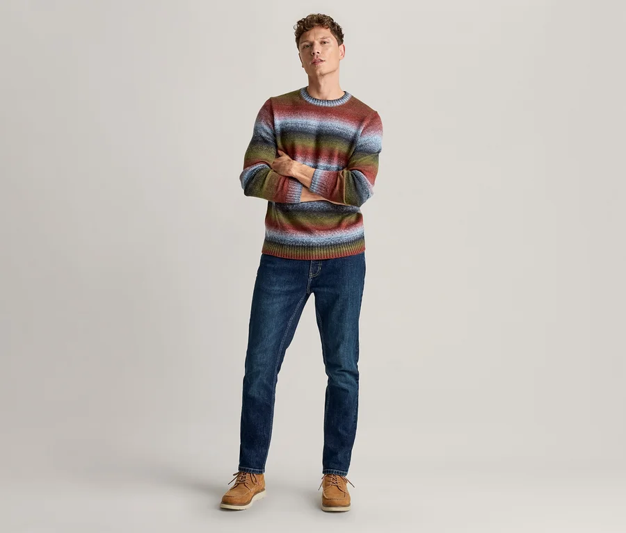 Mann steht mit verschränkten Armen, trägt einen mehrfarbigen Ringelpullover, dunkle Jeans und braune Schnürschuhe.