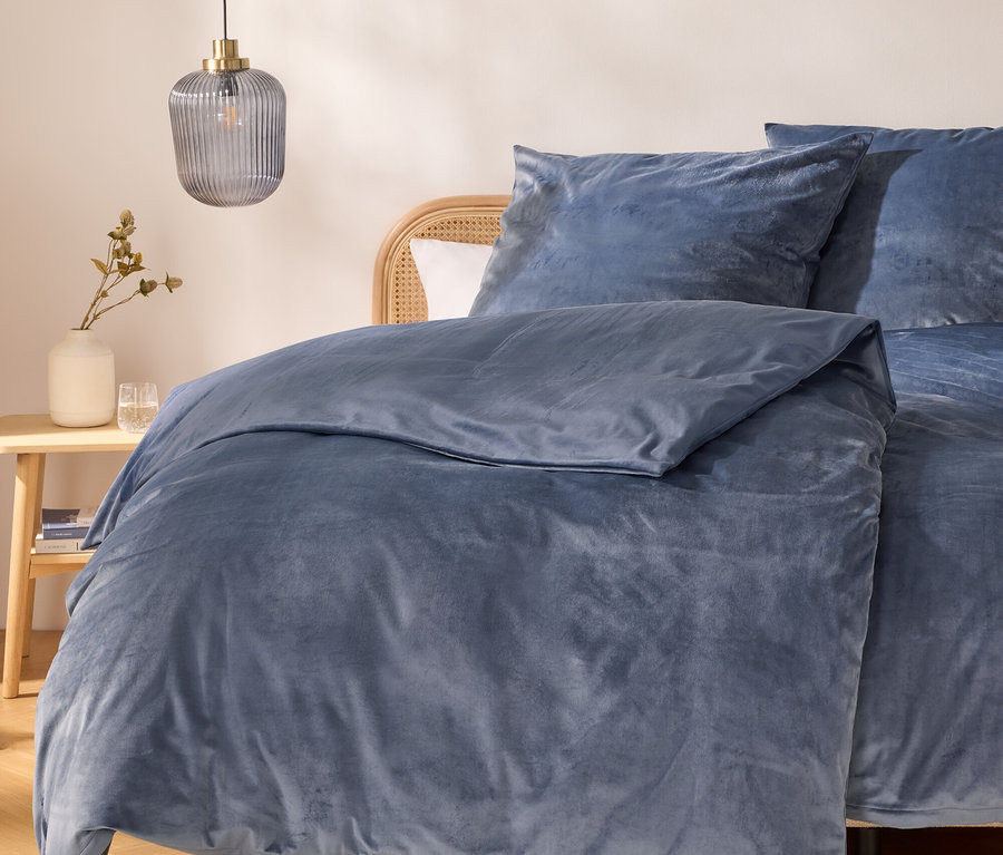Ein Bett mit blauem Samt-Bettbezug und Kissen, einem Rattan-Kopfteil und einem Nachttisch aus Holz mit einer weißen Vase, Trockenpflanzen und zwei Gläsern. Eine gerippte Glas-Pendelleuchte hängt darüber.