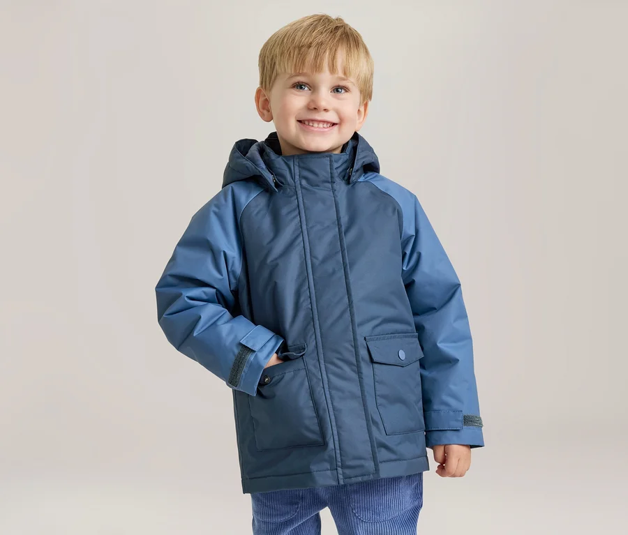 Ein lächelnder kleiner Junge mit blonden Haaren trägt eine zweifarbige blaue Winterjacke, seine linke Hand steckt in der Jackentasche.