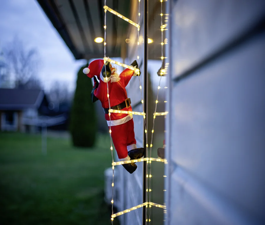 Weihnachtsmann-Figur in rotem Anzug mit weißem Bart klettert an einer Leiter aus warmweißen Lichterketten an der Hauswand in der Dämmerung.