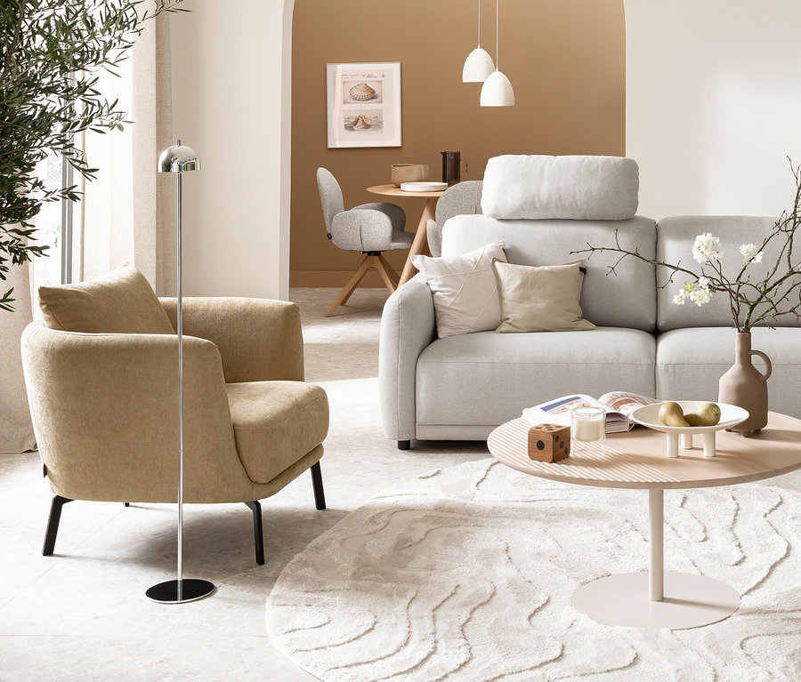 Modernes Wohnzimmer mit hellgrauem Sofa, beigefarbenem Sessel und rundem Couchtisch auf gemustertem Teppich. Daneben steht eine silberfarbene SCHÖNER WOHNEN-Kollektion Akku-Stehleuchte »Bellis«. Im Hintergrund ist ein Essbereich mit Holztisch und grauen Stühlen zu sehen.
