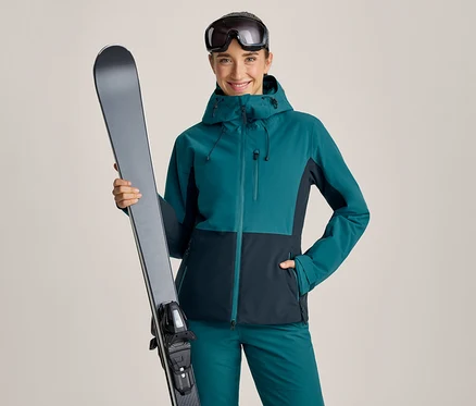Lächelnde Frau in einer tealfarbenen und dunkelblauen Skijacke und passender Hose, die einen grauen Ski vertikal in ihrer linken Hand hält. Eine Skibrille ist auf ihre Stirn geschoben.