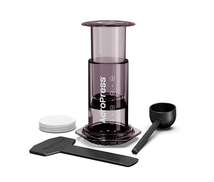 Nahaufnahme eines Aeropress Kaffeebereiters mit Zubehör, inklusive Kaffeelöffel, Rührstab und Papierfilter.