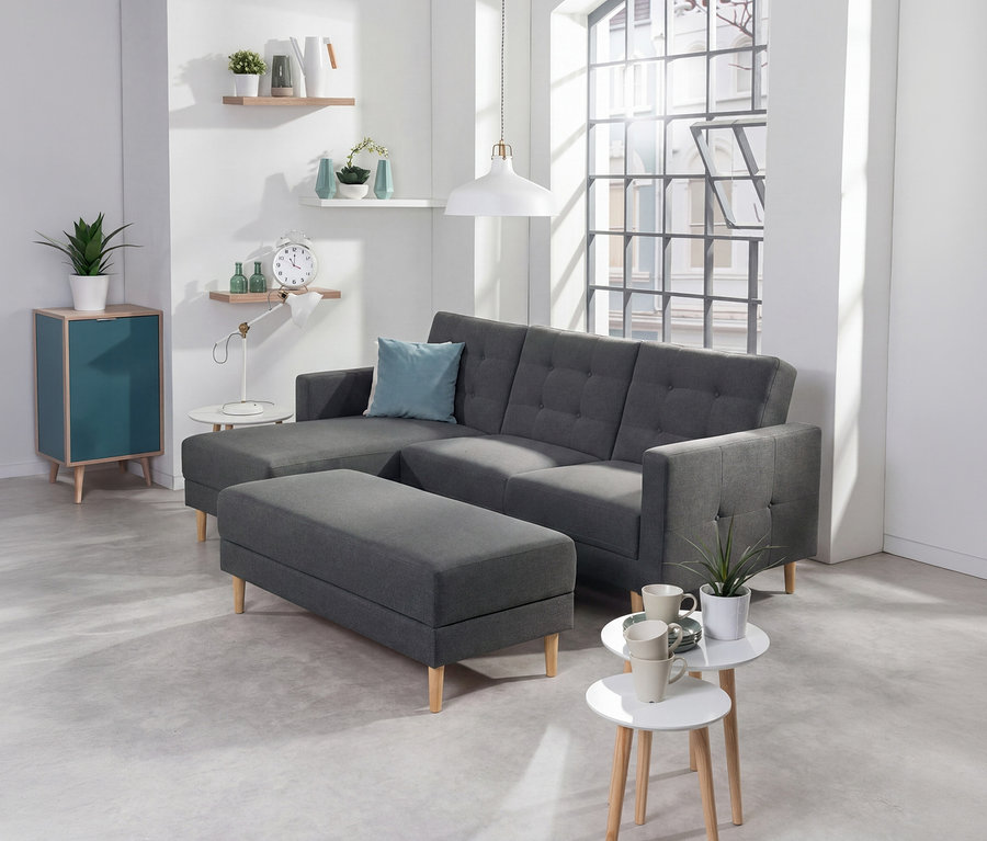 Anthrazitfarbenes Federkern-Eckschlafsofa »Just Relax« mit Stauraumbank in einem modernen Wohnzimmer. Das L-förmige Sofa hat Knopfheftung und ein hellblaues Kissen. Links davon ein türkiser Schrank und Wandregale mit Deko. Davor zwei weißé Beistelltische mit Tassen.