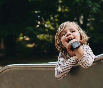 Ein lachendes Kind mit blonden Haaren lehnt sich über den Rand einer Rutsche und trägt eine schwarze Kids Watch Xplora XGO3 2nd Gen.