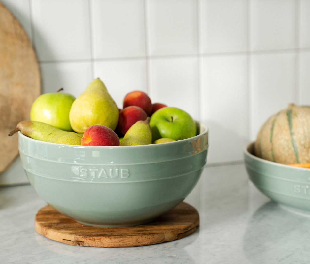 STAUB Keramik-Schüssel in Eukalyptus, gefüllt mit Äpfeln, Birnen und Pfirsichen, auf einem Holzuntersetzer auf einer Marmorarbeitsplatte. Im Hintergrund eine weitere Schüssel mit einer Melone.