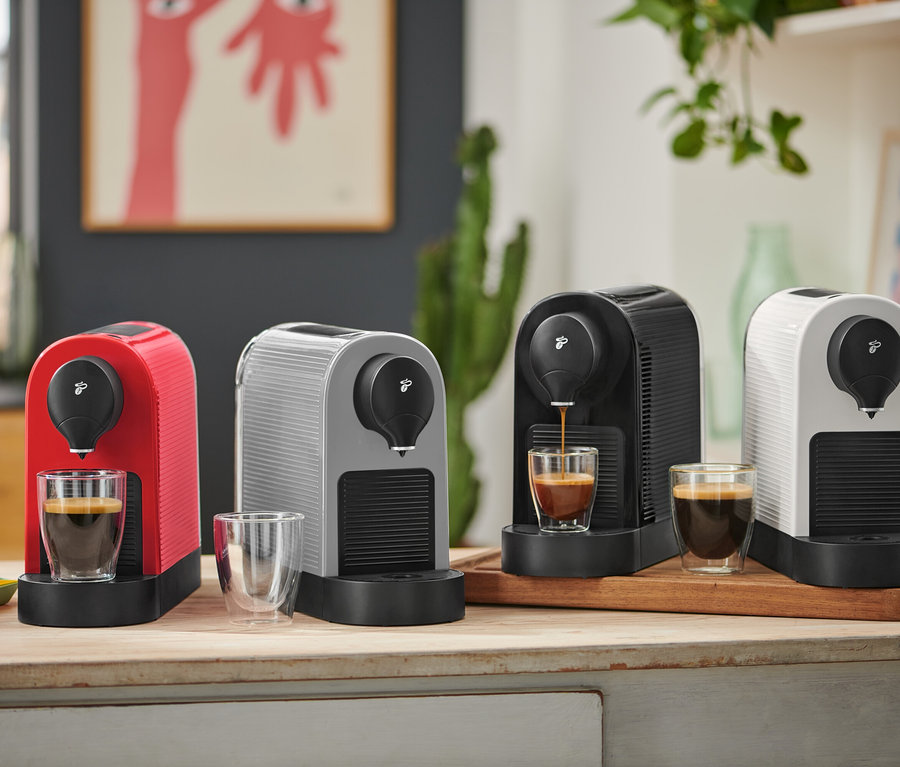 Vier Kaffeemaschinen in Rot, Grau, Schwarz und Weiß stehen auf einer Holztheke. Die schwarze Maschine brüht Kaffee in ein Glas, während die roten und weißen Maschinen Kaffee in ihren Gläsern haben. Vor der grauen Maschine steht ein leeres Glas, und bunte Kaffeekapseln liegen daneben.