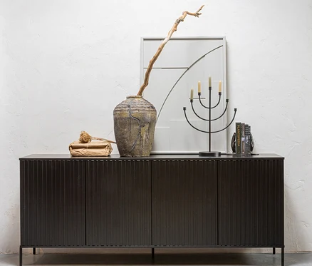 Dunkles geripptes Sideboard mit rustikaler Vase, trockenem Ast, schwarzem fünfarmigem Kerzenständer mit Kerzen und Büchern vor weißer Wand.