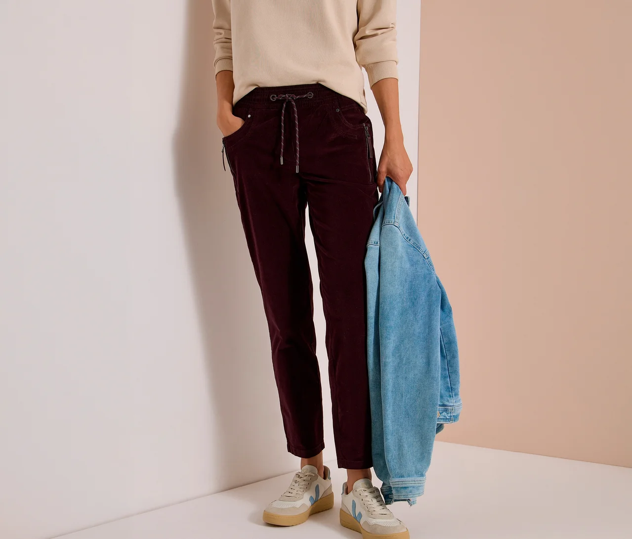 Person trägt beiges Oberteil und bordeauxrote CECIL Cordhose mit Kordelzug und Reißverschlusstaschen. Hält eine Jeansjacke; dazu helle Sneaker.