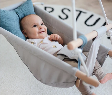 Lächelndes Baby liegt in grauer small foot Babyschaukel »Seaside« mit blauem Kissen.