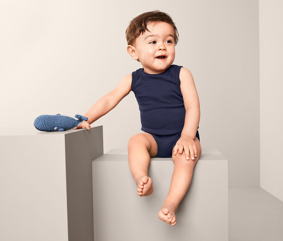 Ein Baby sitzt auf einem grauen Würfel und trägt einen blauen, ärmellosen Body. Neben ihm liegt ein blaues Wal-Spielzeug.