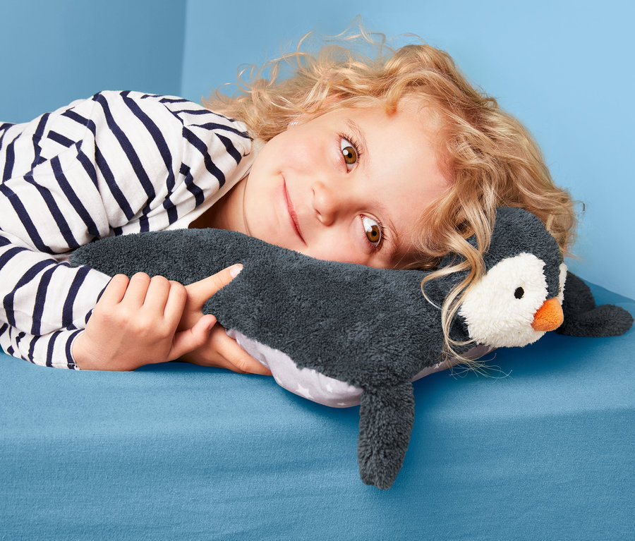 Ein Mädchen liegt auf einem blauen Bett und umarmt ein Kuschelkissen »Pinguin« mit Schlafanzugfach.
