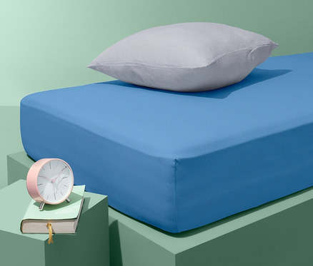 Blaues Jersey-Spannbettlaken auf einem Bett mit einem grauen Kissen. Auf dem Nachttisch liegen ein rosa Wecker und ein Buch.