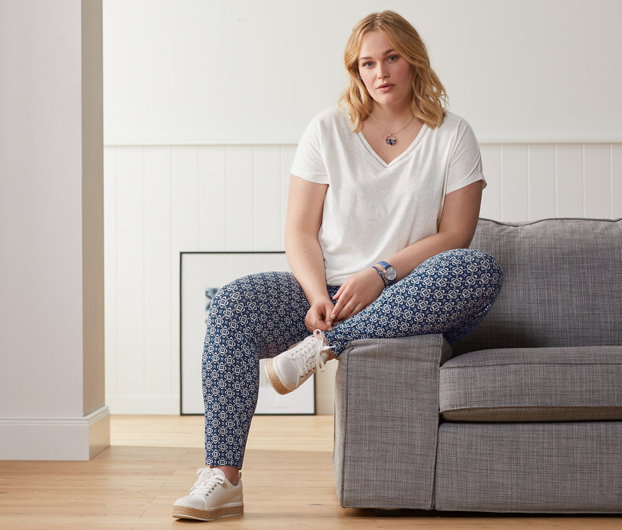 Blonde Frau sitzt auf einem grauen Sofa, trägt ein weißes Shirt aus Leinenmix und eine Jerseyhose mit Alloverprint.