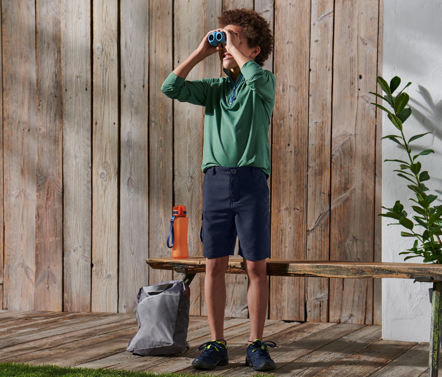 Ein Junge schaut durch ein Fernglas. Er trägt ein grünes Shirt und Funktionsshorts.