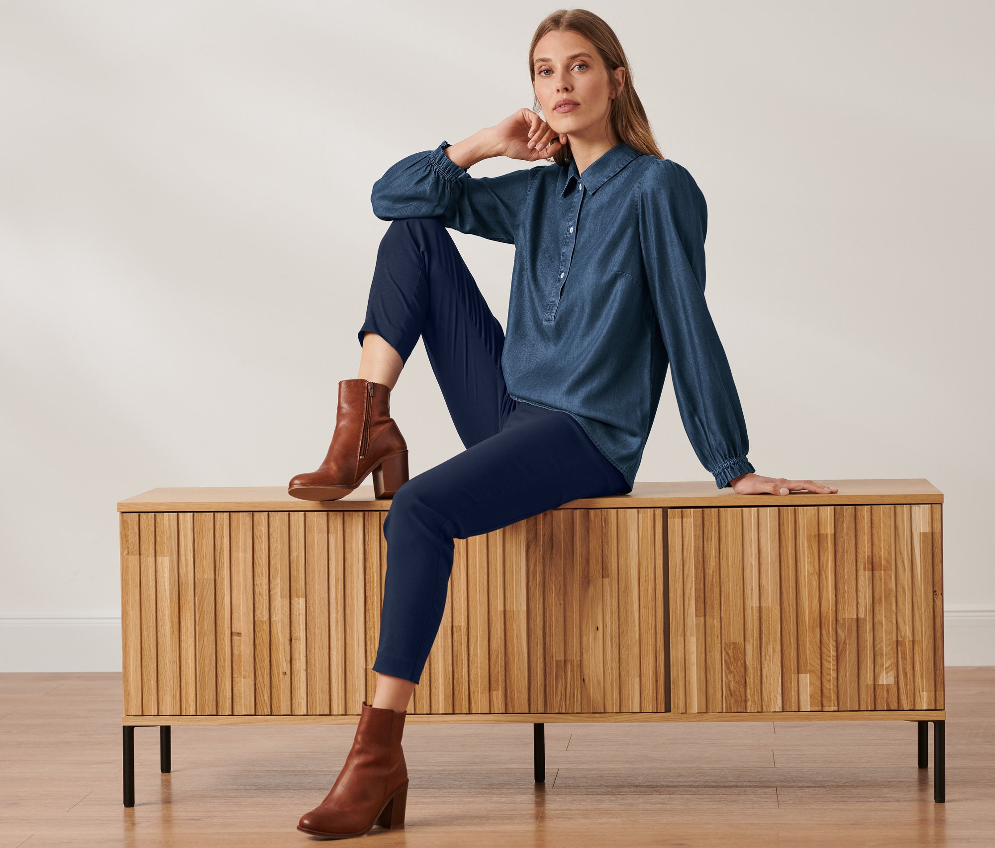 Eine Frau sitzt auf einem Schrank und trägt eine Jeansbluse mit Puffärmeln, dunkelblaue Hosen und braune Stiefel.