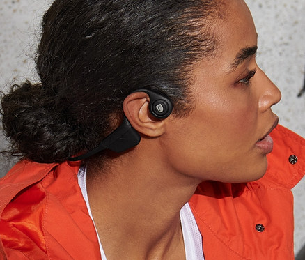 Frau sitzt neben einem Fahrrad und hält einen Helm, sie trägt Bluetooth-Kopfhörer mit Knochenleitung.