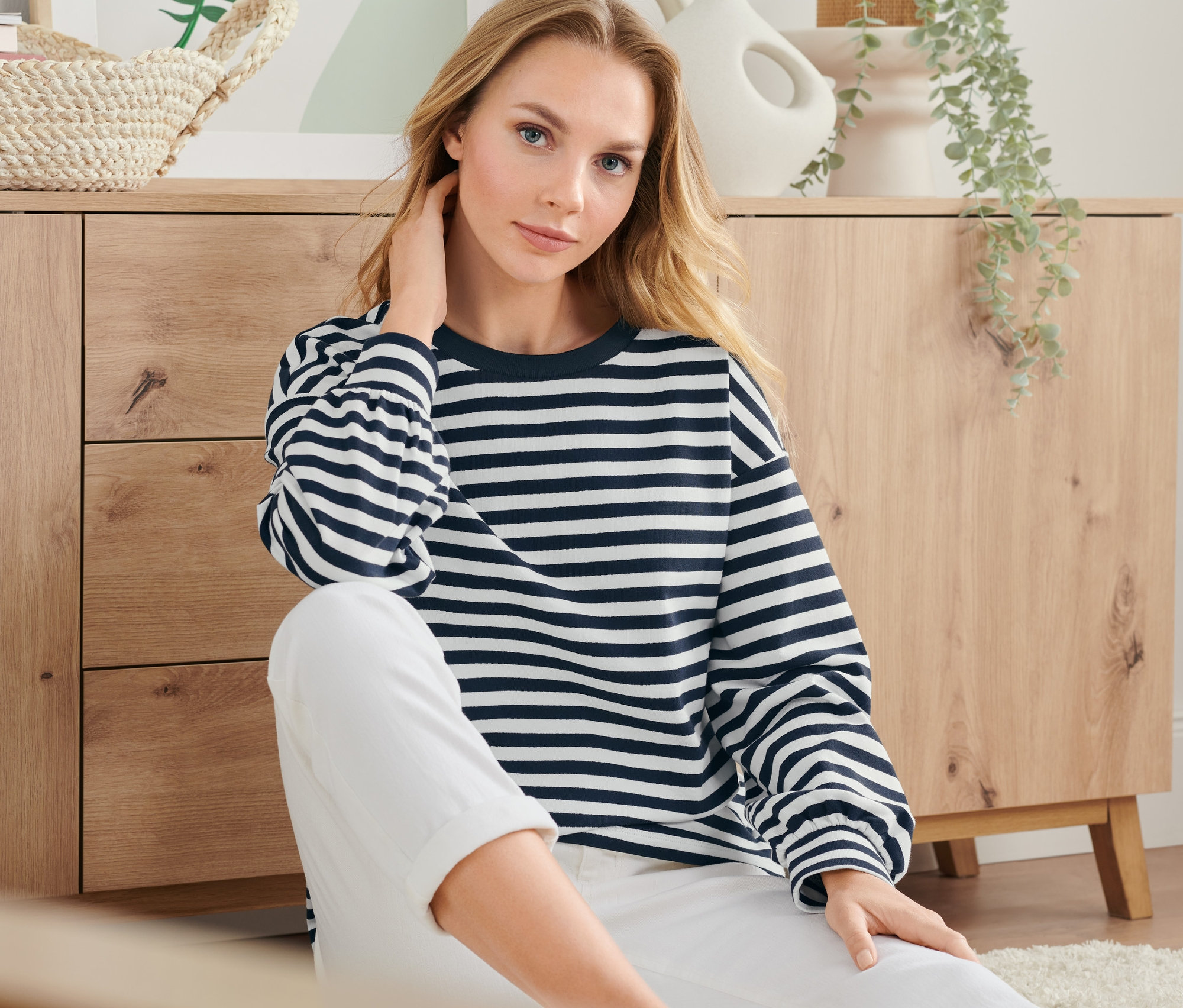 Blonde Frau sitzt auf dem Boden und trägt ein gestreiftes Sweatshirt und weiße Hosen.