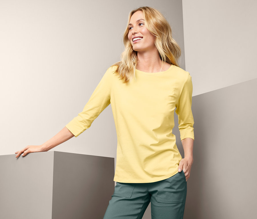 Eine Frau mit hellbraunen Haaren lächelt und lehnt sich an einen grauen Würfel. Sie trägt ein gelbes Shirt mit 3/4-Arm und eine grüne Hose.