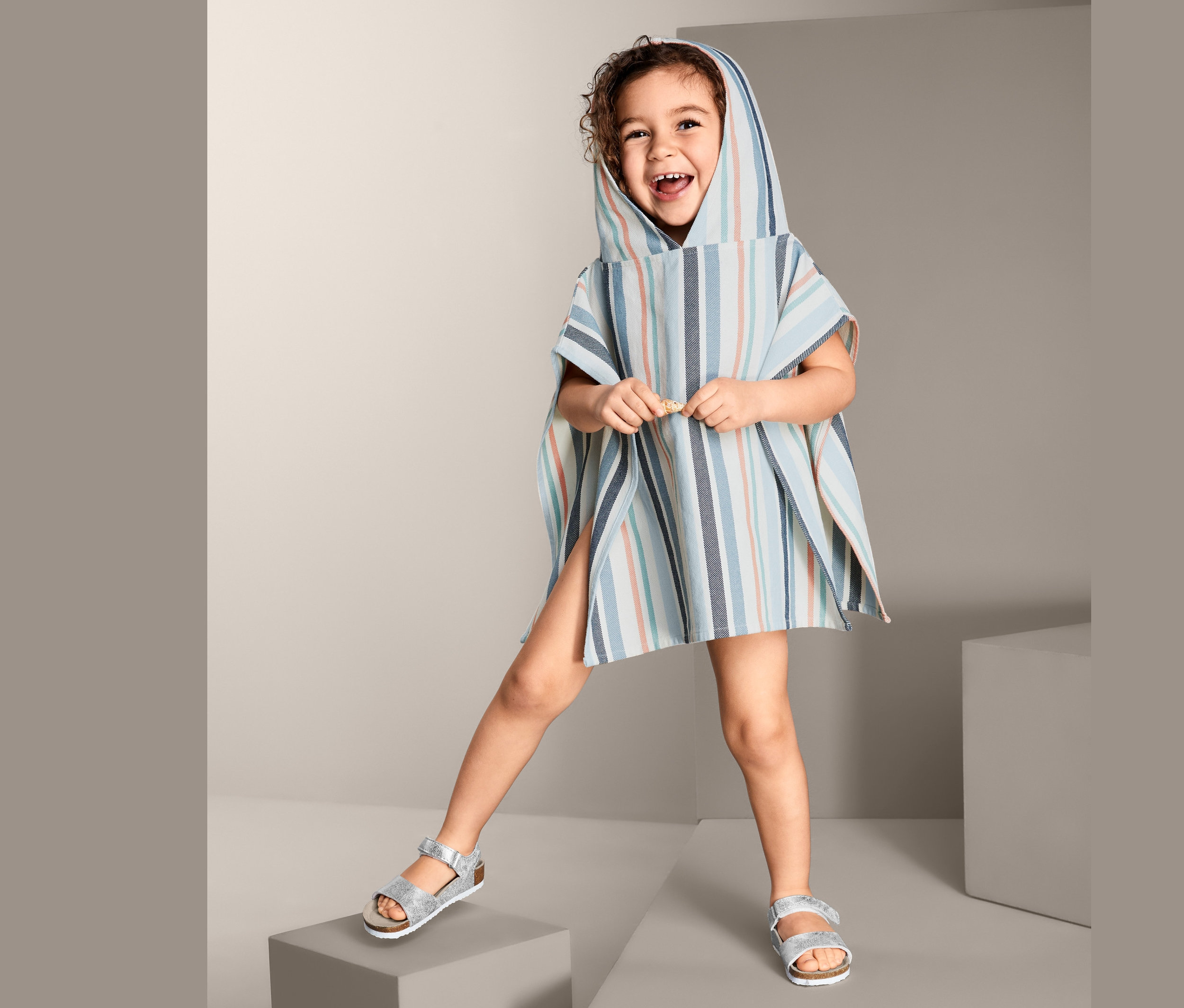 Ein Mädchen steht auf grauen Blöcken und trägt ein Baumwoll-Badecape mit Kapuze und Streifenmuster.