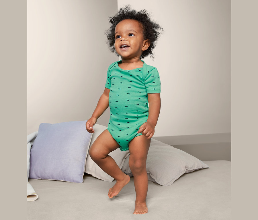 Ein kleiner Junge steht auf einem Bett und trägt einen grünen Baumwollbody mit Fischmuster. Es ist ein Body aus einem 5er-Pack Baby-Baumwollbodys.