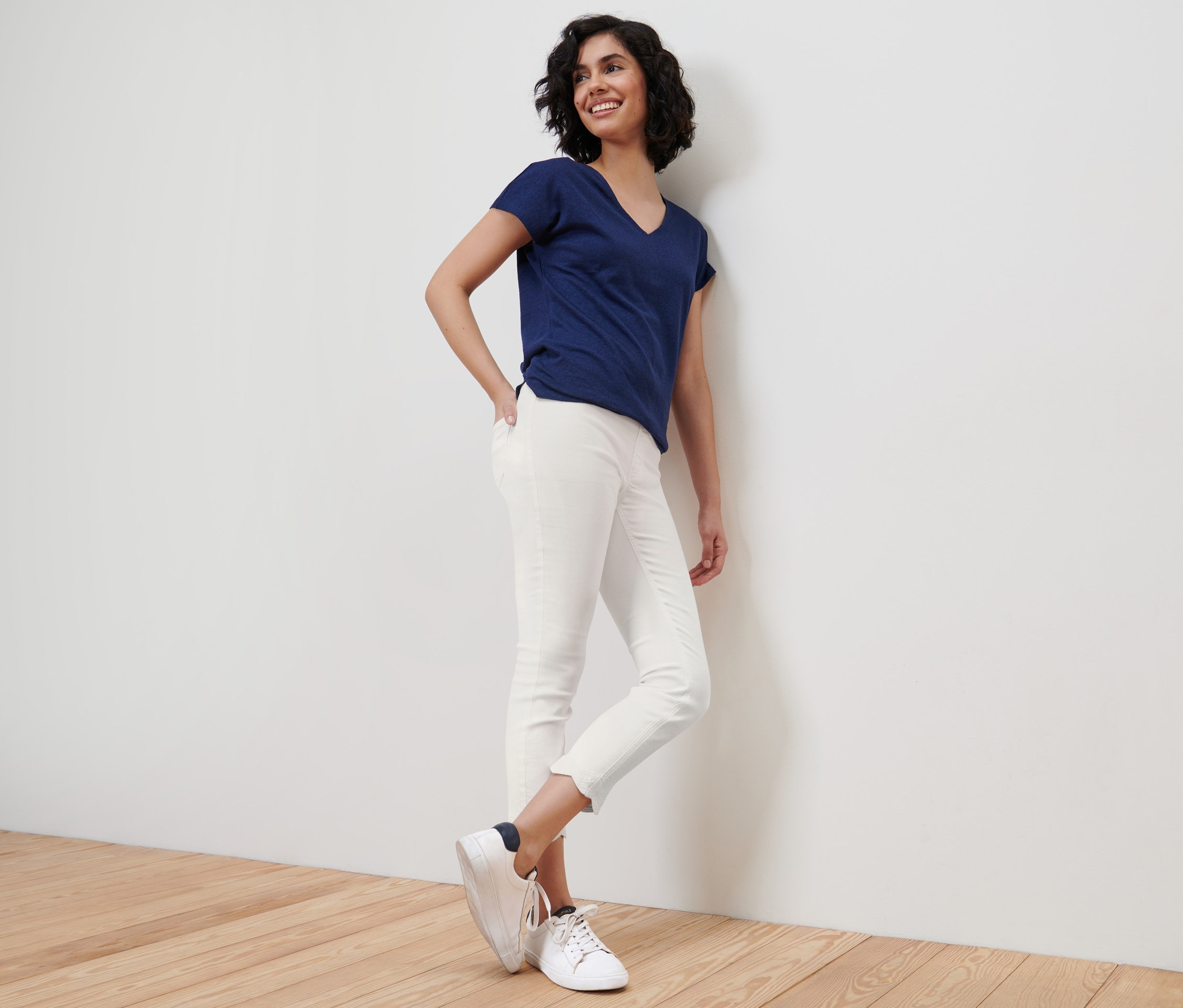 Lächelnde Frau lehnt an einer Wand und trägt ein blaues T-Shirt und weiße 7/8-Treggings mit Lochstickerei.