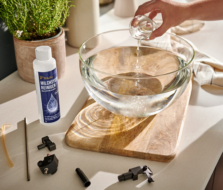 Eine Hand gießt klare Flüssigkeit aus einem Messbecher in eine Glasschüssel mit Wasser. Daneben stehen ein Durgol Milchsystem-Reiniger und Kaffeemaschinenteile.