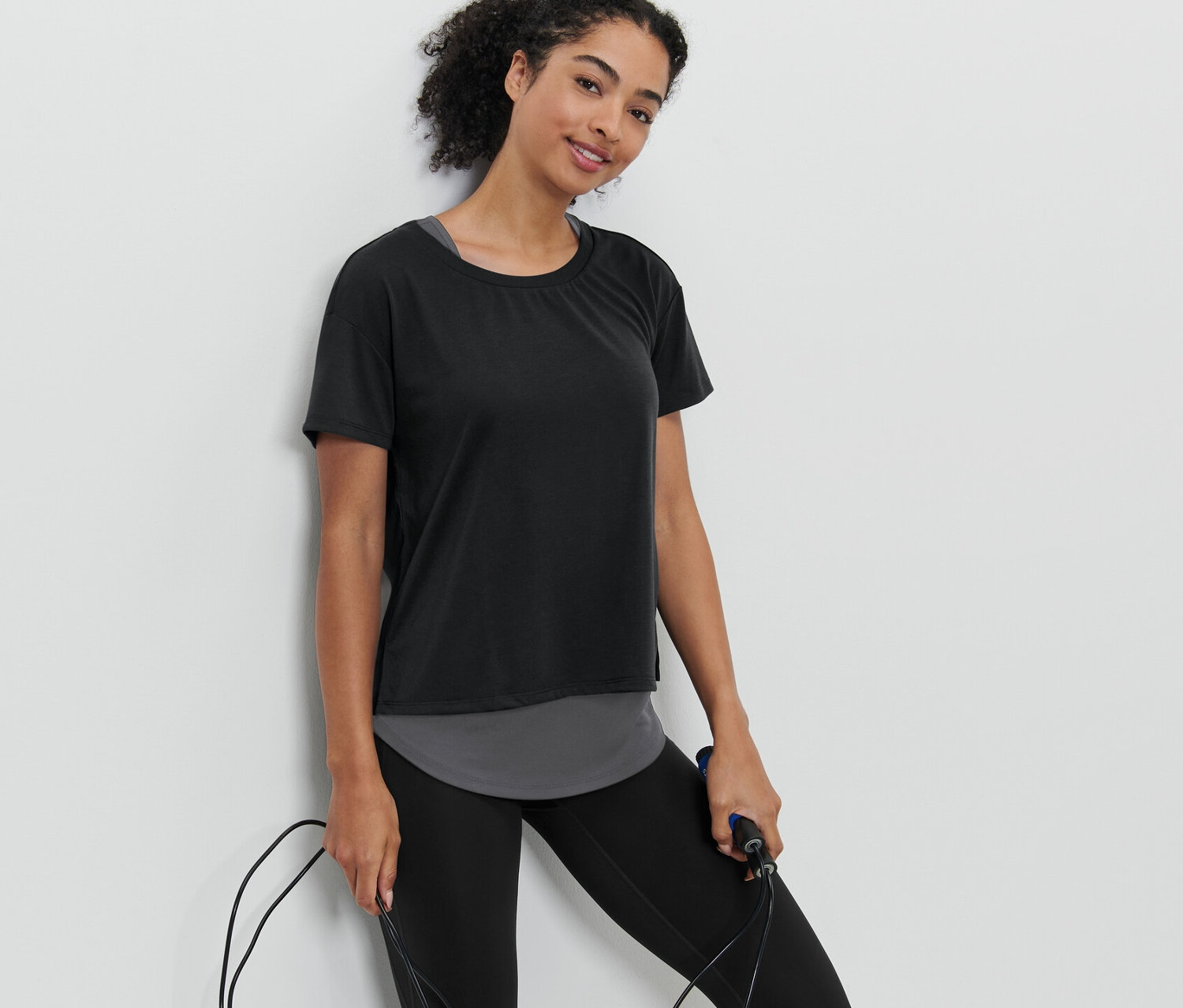 Frau hält ein Springseil und trägt ein schwarzes 2-in-1-Kurzarm-Sportshirt.