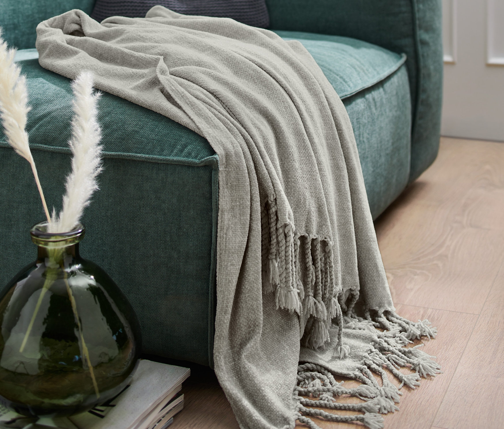 Eine graue Chenille-Wohndecke mit Fransen liegt über einem grünen Sofa.
