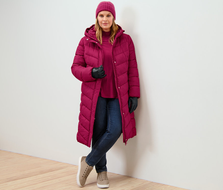 Eine Frau trägt einen himbeerroten Steppmantel mit Kapuze, eine Slim Jeans – Fit »Emma« in Dunkelblau und ein bedrucktes Blusenshirt mit Stehkragen.